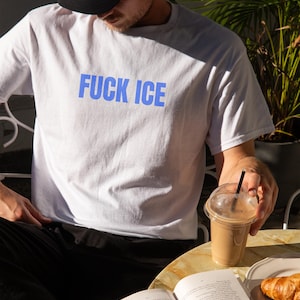 K&ouml;nnte beinhalten: Wei&szlig;es T-Shirt mit dem blauen Text "FUCK ICE". Das Shirt besteht aus einem weichen Material. Das Bild zeigt auch eine schwarze Kappe, ein Getr&auml;nk in einem Plastikbecher, ein Buch und ein Croissant auf einem Teller.