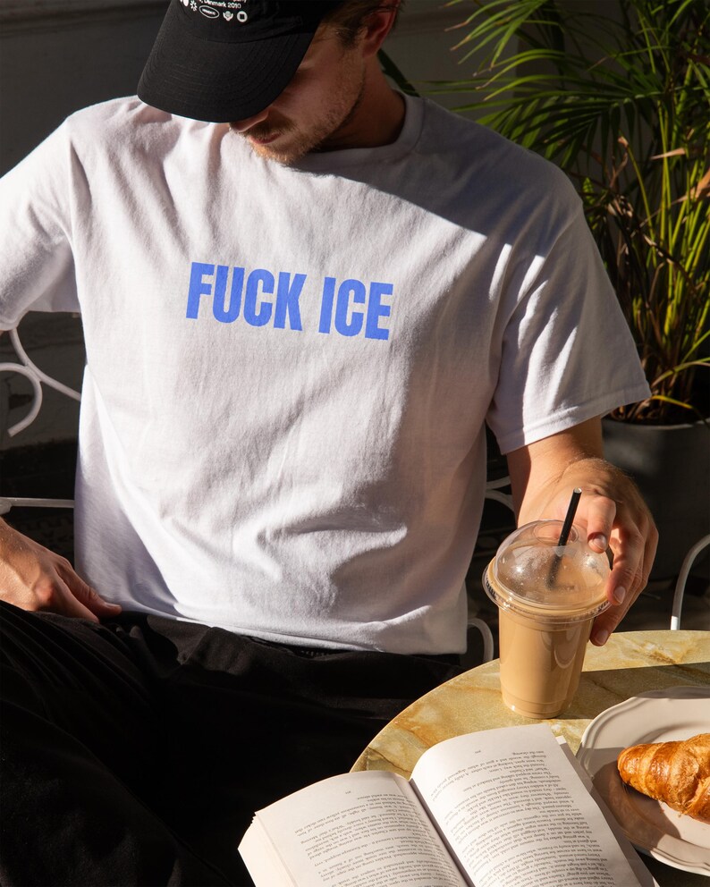 K&ouml;nnte beinhalten: Wei&szlig;es T-Shirt mit dem blauen Text "FUCK ICE". Das Shirt besteht aus einem weichen Material. Das Bild zeigt auch eine schwarze Kappe, ein Getr&auml;nk in einem Plastikbecher, ein Buch und ein Croissant auf einem Teller.