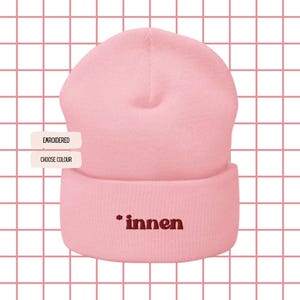 Peut inclure: Un bonnet rose clair avec un revers. Le mot "innen" est brodé en rouge foncé sur le revers. Le fond est un motif quadrillé de lignes roses sur fond blanc. Les textes "EMBROIDERED" et "CHOOSE COLOUR" sont visibles.