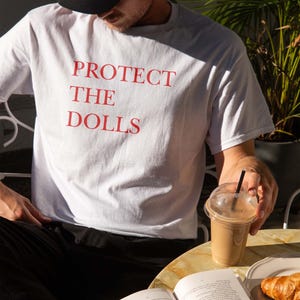 May include: White t-shirt with the red text "PROTECT THE DOLLS". The person is wearing a black baseball cap. A clear plastic cup with a brown beverage and a black straw is on the table. A book and a croissant are also on the table.