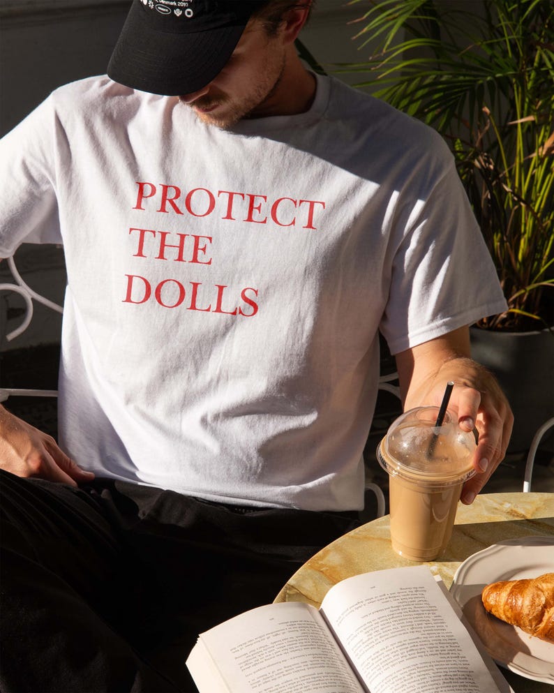 May include: White t-shirt with the red text "PROTECT THE DOLLS". The person is wearing a black baseball cap. A clear plastic cup with a brown beverage and a black straw is on the table. A book and a croissant are also on the table.