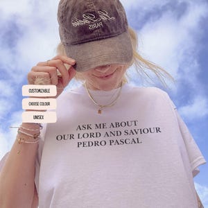 May include: White t-shirt with the text "ASK ME ABOUT OUR LORD AND SAVIOUR PEDRO PASCAL" in black. A brown baseball cap with white text is worn. The background is a blue sky with clouds.