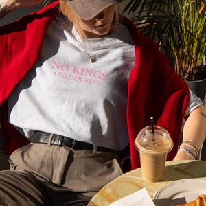 Op de afbeelding: Lichtgrijs t-shirt met de roze tekst "NO KINGS ONLY QUEENS". De persoon draagt een bruine baseballpet en een rode trui. Een koffie en een croissant staan op tafel.