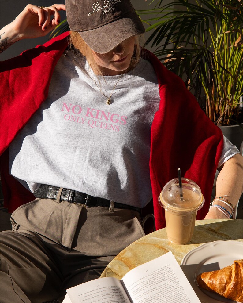 Op de afbeelding: Lichtgrijs t-shirt met de roze tekst "NO KINGS ONLY QUEENS". De persoon draagt een bruine baseballpet en een rode trui. Een koffie en een croissant staan op tafel.