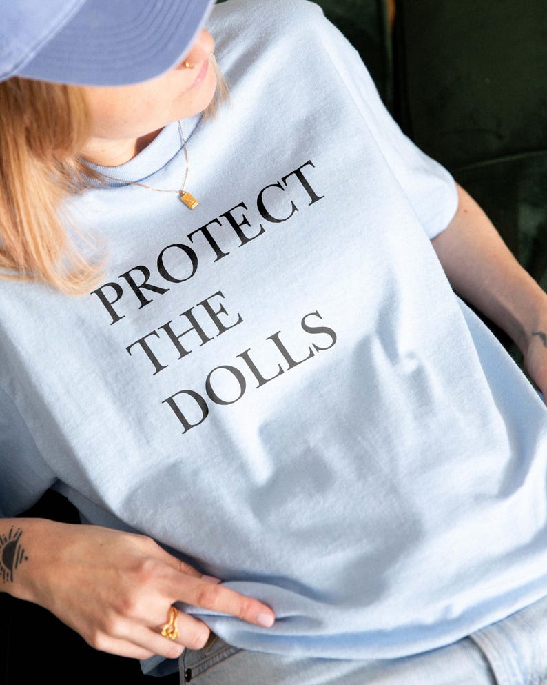 May include: Light blue t-shirt with the text "PROTECT THE DOLLS" in black. The shirt is made of a soft material. The person is wearing a blue cap and a gold necklace.