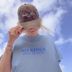 Op de afbeelding: Een lichtblauw T-shirt met de blauwe tekst "NO KINGS ONLY QUEENS". Een bruine baseballcap met witte cursieve tekst wordt gedragen. De achtergrond is een blauwe lucht met witte wolken.