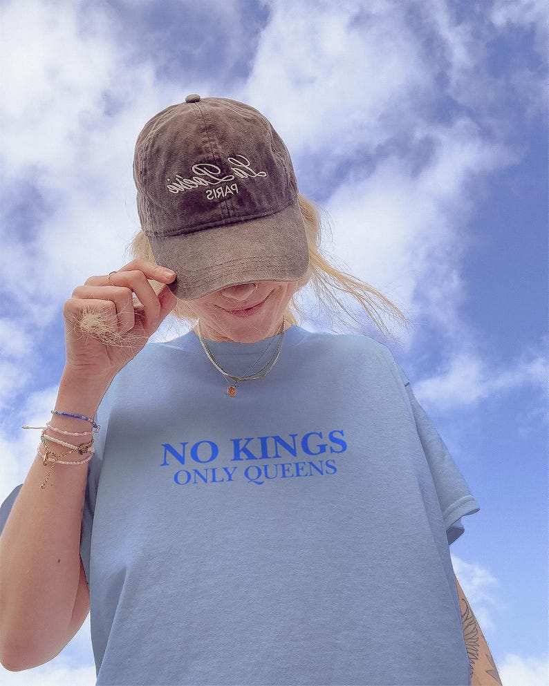 Op de afbeelding: Een lichtblauw T-shirt met de blauwe tekst "NO KINGS ONLY QUEENS". Een bruine baseballcap met witte cursieve tekst wordt gedragen. De achtergrond is een blauwe lucht met witte wolken.