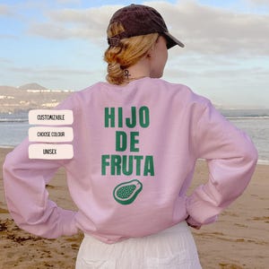 Op de afbeelding: Een lichtroze sweatshirt met de tekst "HIJO DE FRUTA" in het groen, samen met een groene papaja-illustratie. Het sweatshirt heeft ook de woorden "CUSTOMIZABLE", "CHOOSE COLOUR" en "UNISEX" aan de linkerkant. Een persoon draagt het sweatshirt.