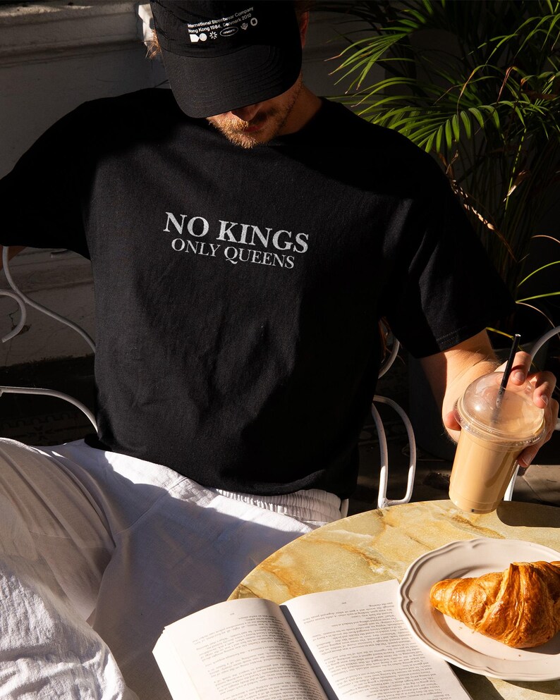 Op de afbeelding: Zwart t-shirt met de witte tekst "NO KINGS ONLY QUEENS". De persoon draagt een zwarte pet en een witte broek. Een croissant ligt op een bord naast een open boek en een drankje.