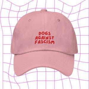 Może przedstawiać: R&oacute;żowa czapka z daszkiem z haftem "DOGS AGAINST FASCISM" w kolorze czerwonym.