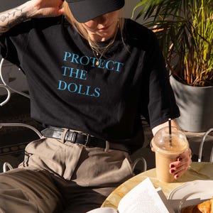 May include: Black t-shirt with the text "PROTECT THE DOLLS" in blue. The person is wearing a black cap and tan pants. A cup of coffee, a book, and a croissant are on the table.