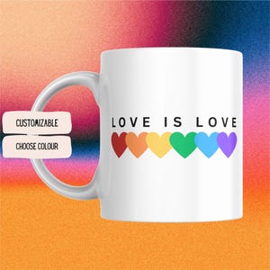 May include: White ceramic mug with the text "LOVE IS LOVE" and a row of rainbow-colored hearts. The mug has a curved handle and the words "CUSTOMIZABLE" and "CHOOSE COLOUR" are on a separate label.