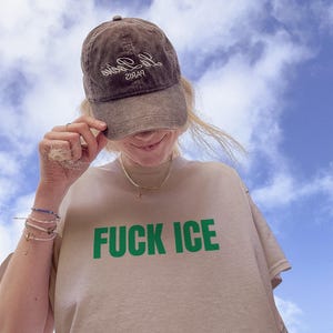 K&ouml;nnte beinhalten: Braune Baseballkappe mit wei&szlig;em Text und beiges T-Shirt mit den gr&uuml;nen Worten "FUCK ICE". Der Himmel ist blau mit wei&szlig;en Wolken.