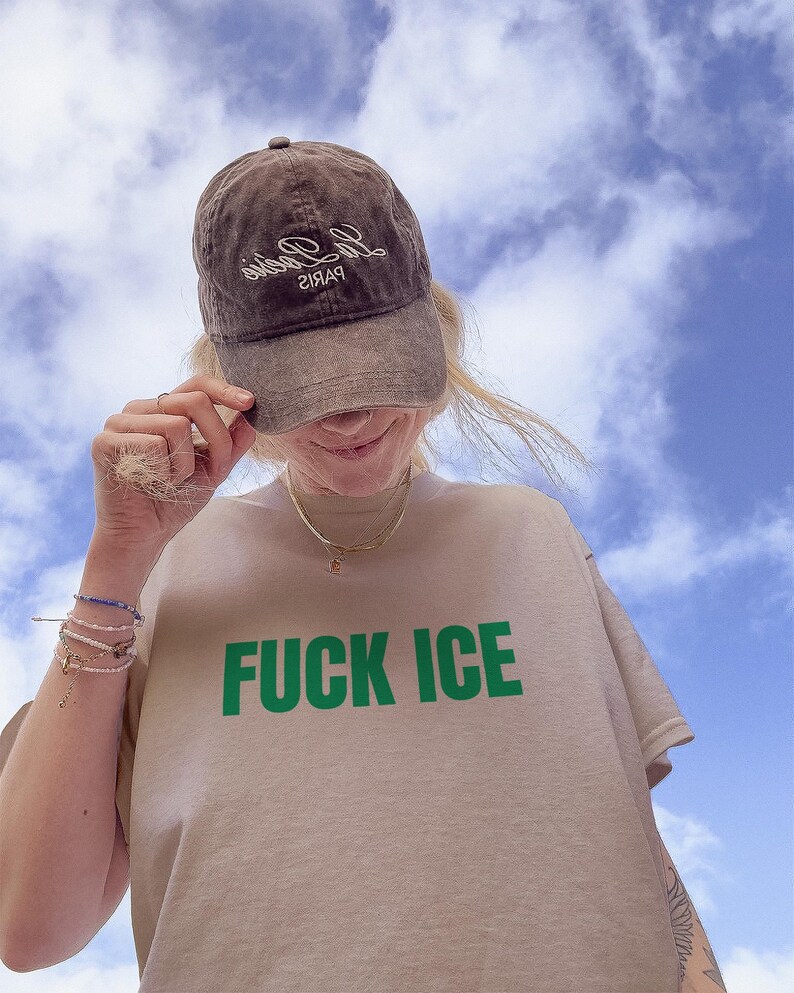 K&ouml;nnte beinhalten: Braune Baseballkappe mit wei&szlig;em Text und beiges T-Shirt mit den gr&uuml;nen Worten "FUCK ICE". Der Himmel ist blau mit wei&szlig;en Wolken.