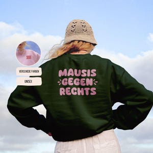 May include: A dark green unisex sweatshirt with the text "MAUSIS GEGEN RECHTS" in pink, centered on the back. The image includes a small inset showing a pink sweatshirt with the same text. The words "VERSCHIEDE FARBEN" and "UNISEX" are also visible.