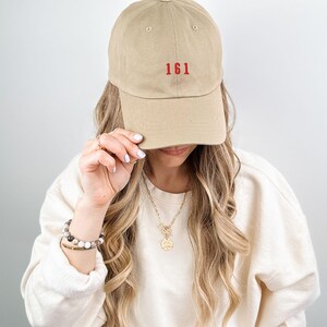 May include: A tan baseball cap with red embroidered text that reads "161". The cap is being worn by a person with long blonde hair.
