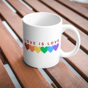 May include: White ceramic mug with the text "LOVE IS LOVE" in red, above a row of colorful hearts. The hearts are in the colors of the rainbow. The mug has a curved handle and is sitting on a wooden surface.