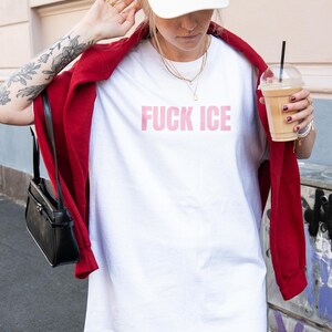 K&ouml;nnte beinhalten: Wei&szlig;es T-Shirt mit dem rosa Text "FUCK ICE". Die Person tr&auml;gt eine rote Jacke, eine wei&szlig;e Kappe und h&auml;lt ein Kaffeegetr&auml;nk. Eine schwarze Handtasche h&auml;ngt an der Schulter.