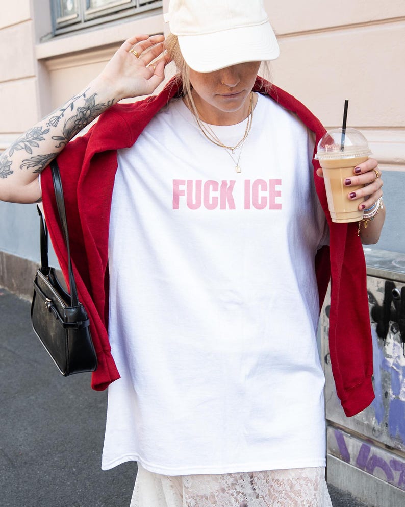 K&ouml;nnte beinhalten: Wei&szlig;es T-Shirt mit dem rosa Text "FUCK ICE". Die Person tr&auml;gt eine rote Jacke, eine wei&szlig;e Kappe und h&auml;lt ein Kaffeegetr&auml;nk. Eine schwarze Handtasche h&auml;ngt an der Schulter.