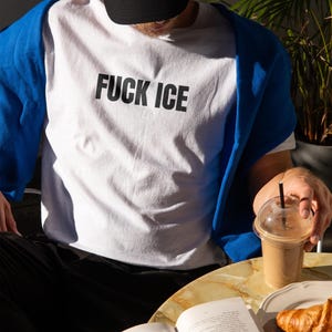 K&ouml;nnte beinhalten: Wei&szlig;es T-Shirt mit dem Text "FUCK ICE" in schwarzen Blockbuchstaben. Ein blauer Pullover ist &uuml;ber die Schultern gelegt. Eine schwarze Kappe wird getragen. Eine Person h&auml;lt ein Getr&auml;nk mit Strohhalm und ein Buch ist auf einem Tisch ge&ouml;ffnet.