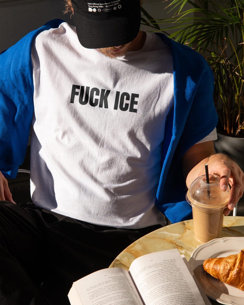 K&ouml;nnte beinhalten: Wei&szlig;es T-Shirt mit dem Text "FUCK ICE" in schwarzen Blockbuchstaben. Ein blauer Pullover ist &uuml;ber die Schultern gelegt. Eine schwarze Kappe wird getragen. Eine Person h&auml;lt ein Getr&auml;nk mit Strohhalm und ein Buch ist auf einem Tisch ge&ouml;ffnet.