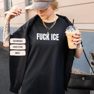 K&ouml;nnte beinhalten: Schwarzes Unisex-Sweatshirt mit dem Text "FUCK ICE" in wei&szlig;en Blockbuchstaben. Das Bild enth&auml;lt auch drei wei&szlig;e rechteckige Etiketten mit der Aufschrift "CUSTOMIZABLE", "CHOOSE COLOUR" und "UNISEX".