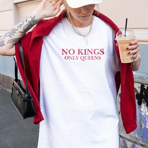 Op de afbeelding: Een wit t-shirt met de tekst "NO KINGS ONLY QUEENS" in rode letters. De persoon draagt een rode jas, een witte pet en houdt een drankje vast in een doorzichtige plastic beker met een zwart rietje. Een zwarte handtas hangt aan de schouder.