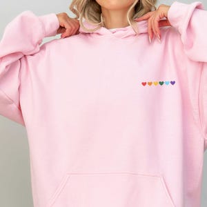 Può includere: Una felpa con cappuccio rosa con una fila di cuori arcobaleno ricamata sul davanti.