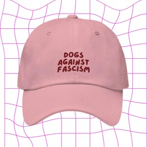 Może przedstawiać: R&oacute;żowa czapka z daszkiem z haftowanym napisem "DOGS AGAINST FASCISM" w kolorze czerwonym.