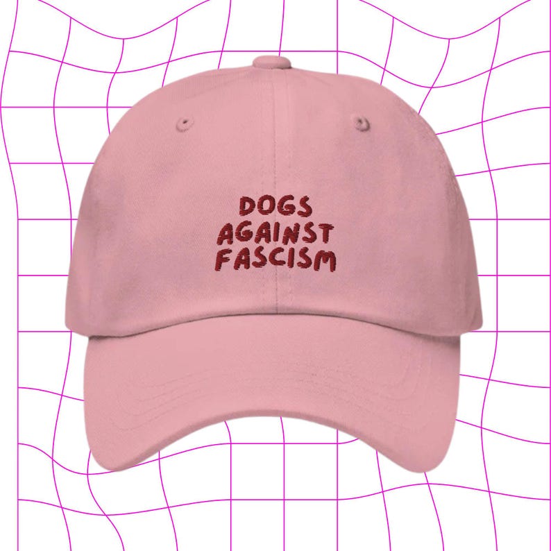 Może przedstawiać: R&oacute;żowa czapka z daszkiem z haftowanym napisem "DOGS AGAINST FASCISM" w kolorze czerwonym.