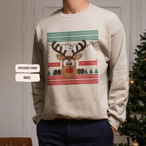 Könnte beinhalten: Beiges Unisex-Weihnachtssweatshirt mit einem Rentier-Design und dem Schriftzug "MERRY CHRISTMAS". Das Rentier hat eine rote Nase und ist mit Lichterketten geschmückt. Das Sweatshirt hat grüne und rote gemusterte Ränder.