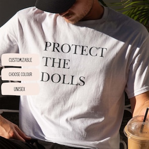 May include: White t-shirt with the text "PROTECT THE DOLLS" in black. The shirt also has the words "CUSTOMIZABLE", "CHOOSE COLOUR", and "UNISEX" in a white box. The shirt is a casual, unisex garment.