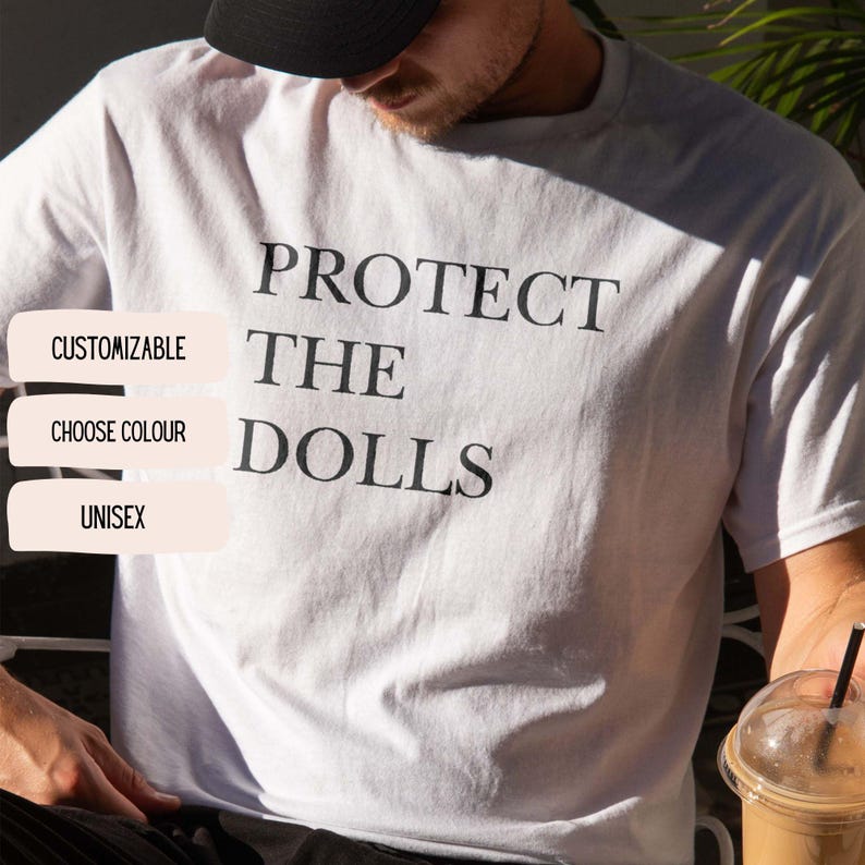 May include: White t-shirt with the text "PROTECT THE DOLLS" in black. The shirt also has the words "CUSTOMIZABLE", "CHOOSE COLOUR", and "UNISEX" in a white box. The shirt is a casual, unisex garment.