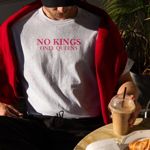 Op de afbeelding: Lichtgrijs t-shirt met de roze tekst "NO KINGS ONLY QUEENS". De persoon draagt een zwarte pet en een rode trui. Een koffie en een croissant liggen op tafel.