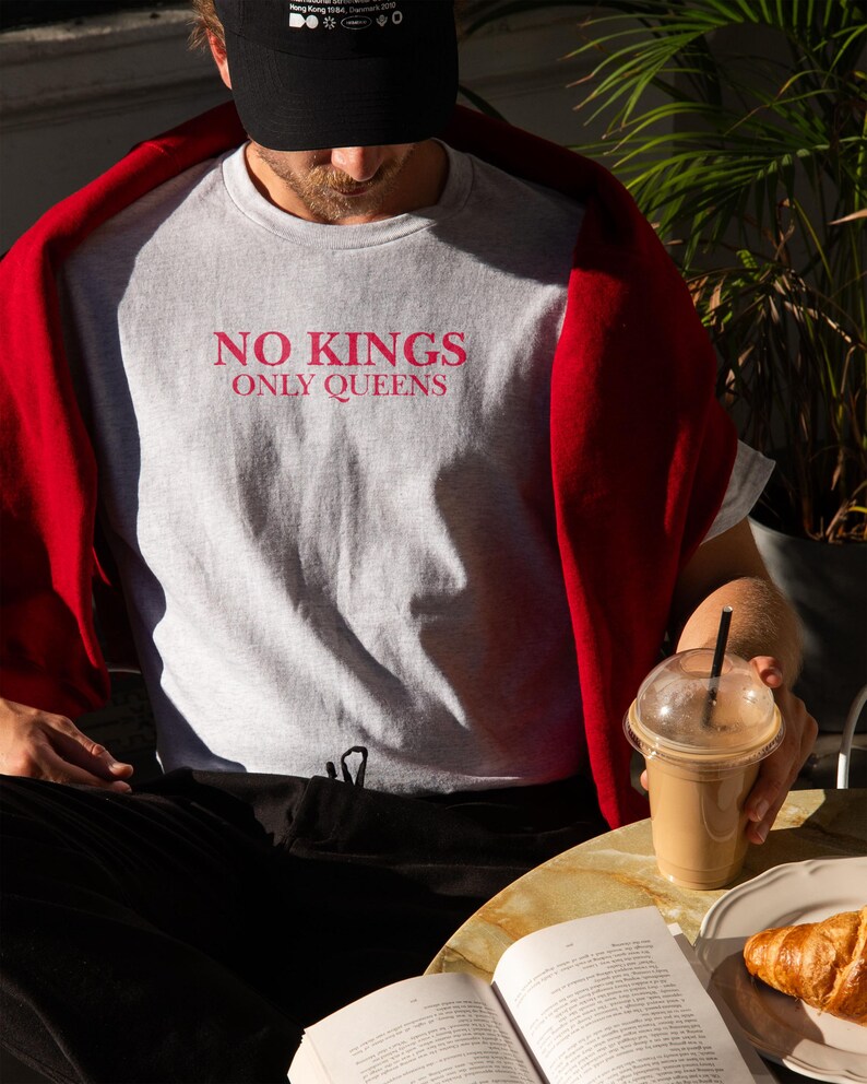 Op de afbeelding: Lichtgrijs t-shirt met de roze tekst "NO KINGS ONLY QUEENS". De persoon draagt een zwarte pet en een rode trui. Een koffie en een croissant liggen op tafel.