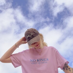 Op de afbeelding: Een roze t-shirt met de tekst "NO KINGS ONLY QUEENS" in lichtblauw. De persoon draagt een bruine baseballcap met een logo en een gouden ketting. De achtergrond is een blauwe lucht met witte wolken.