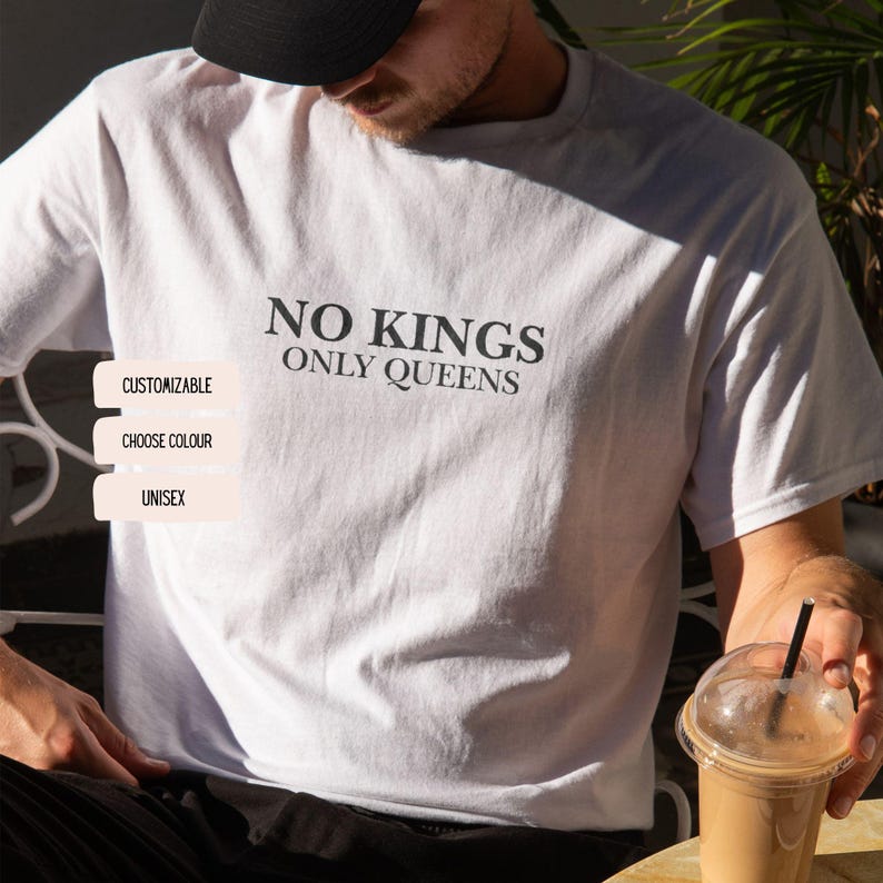 Op de afbeelding: Wit unisex T-shirt met de zwarte tekst "NO KINGS ONLY QUEENS". Het shirt is aanpasbaar en verkrijgbaar in verschillende kleuren. Het model draagt een zwarte pet en houdt een drankje vast.