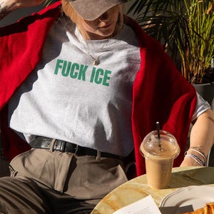 K&ouml;nnte beinhalten: Ein hellgraues T-Shirt mit den Worten "FUCK ICE" in gr&uuml;nen Blockbuchstaben. Die Person tr&auml;gt eine braune Baseballkappe und einen roten Pullover. Ein Kaffeegetr&auml;nk und ein Croissant liegen auf dem Tisch.