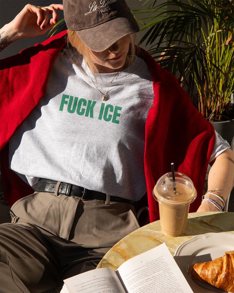 K&ouml;nnte beinhalten: Ein hellgraues T-Shirt mit den Worten "FUCK ICE" in gr&uuml;nen Blockbuchstaben. Die Person tr&auml;gt eine braune Baseballkappe und einen roten Pullover. Ein Kaffeegetr&auml;nk und ein Croissant liegen auf dem Tisch.