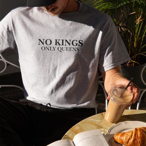 Op de afbeelding: Lichtgrijs t-shirt met de zwarte tekst "NO KINGS ONLY QUEENS". De afbeelding toont ook een zwarte baseball cap, een kopje koffie, een croissant en een open boek op een tafel.