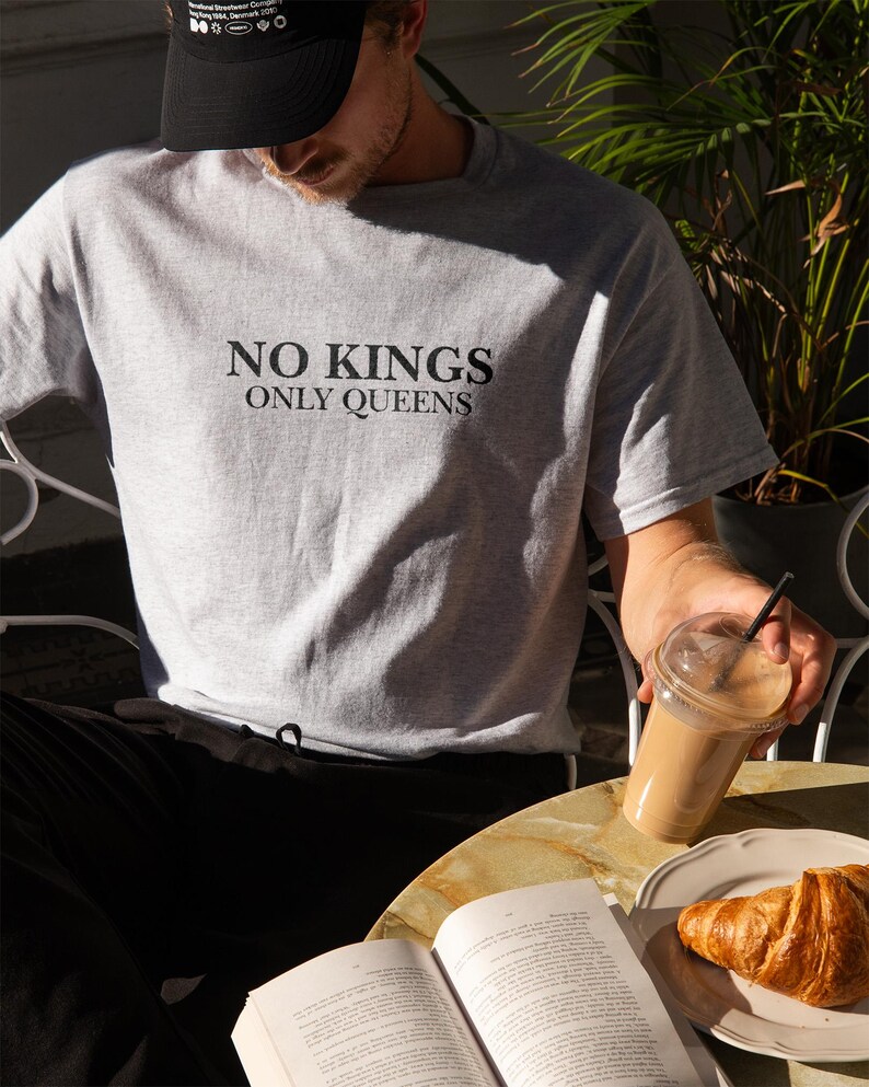 Op de afbeelding: Lichtgrijs t-shirt met de zwarte tekst "NO KINGS ONLY QUEENS". De afbeelding toont ook een zwarte baseball cap, een kopje koffie, een croissant en een open boek op een tafel.