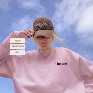 Puede incluir: Una persona con una gorra de béisbol marrón con "París" bordado y una sudadera rosa claro con la palabra "innen" impresa. La imagen también muestra las palabras "BESTICKT", "VERSCHIEDE FARBEN" y "UNISEX".
