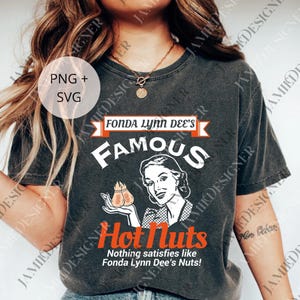 May include: Dark gray t-shirt featuring the text "FONDA LYNN DEE'S FAMOUS Hot Nuts" with a vintage-style illustration of a woman holding nuts. The shirt also includes the phrase "Nothing satisfies like Fonda Lynn Dee's Nuts!"