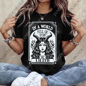 May include: Black t-shirt featuring a white graphic of a woman with horns, curly hair, and the text "IN A WORLD FULL OF EVES BE LILITH". The design is framed in a rectangular border. The shirt is a short-sleeved crew neck.