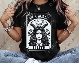 May include: Black t-shirt featuring a white graphic of a woman with horns, curly hair, and the text "IN A WORLD FULL OF EVES BE LILITH". The design is framed in a rectangular border. The shirt is a short-sleeved crew neck.
