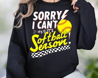 May include: Black sweatshirt with the text "SORRY I CAN'T IT'S Softball Season" in white, red, and yellow. A yellow softball with red stitching is on the right. A checkered pattern is at the bottom.