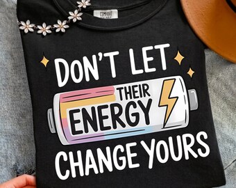 May include: A black t-shirt featuring a graphic of a battery with a lightning bolt icon. The text reads "Don't Let Their Energy Change Yours." The design includes pastel-colored accents within the battery graphic and small yellow star shapes.