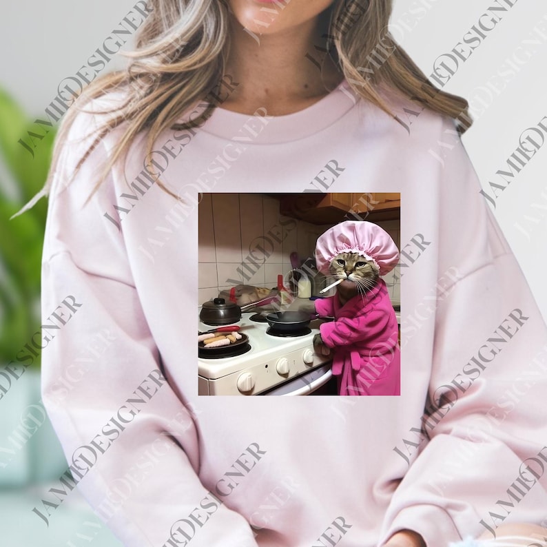 A woman wearing a pink sweater with a printed image of a cat wearing a pink hat and robe, standing in a kitchen and cooking on a stove.