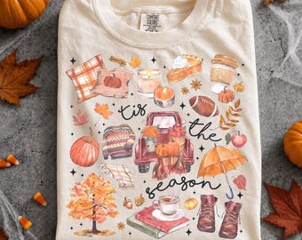 Puede incluir: Una camiseta de color crema con un estampado gráfico de temática otoñal y el texto 'tis the season'. El diseño incluye ilustraciones de un camión rojo, calabazas, un suéter, un trozo de tarta, una taza de café, un paraguas y botas.