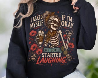 May include: A black long-sleeve sweatshirt featuring a graphic of a skeleton wearing sunglasses, a leopard print headband, and a scarf. The skeleton holds a drink, surrounded by red flowers and the text: I ASKED MYSELF IF I'M OKAY AND WE BOTH STARTED LAUGHING.